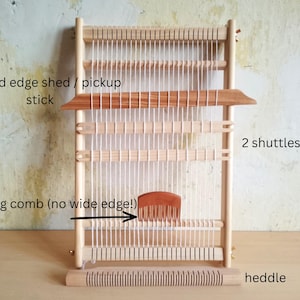 May include: A wooden weaving loom with a beveled edge shed/pickup stick, two shuttles, a weaving comb, and a heddle. The loom is made of light-colored wood and has white warp threads strung vertically. The weaving comb is brown.