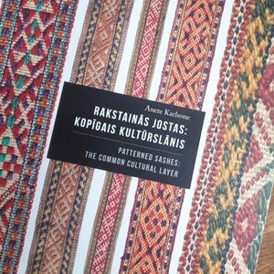 Puede incluir: Un libro titulado "Rakstainās Jostas: Kopīgais Kultūraslānis" de Anete Karlsone, con diseños de fajas estampadas coloridas en rojo, verde y marrón. La portada tiene una etiqueta negra con texto blanco y está sobre una superficie de madera.