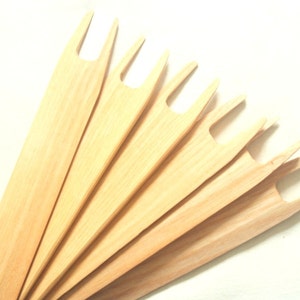 Puede incluir: Un juego de siete agujas de tejer de madera con un extremo bifurcado. Las agujas están dispuestas en forma de abanico y son de madera marrón claro.