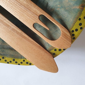 May include: Two wooden weaving tools, a shuttle and a pick, are displayed on a painted surface. The shuttle is long and flat with a pointed end. The pick has a U-shaped opening. The background is a mix of blue, green, and yellow.