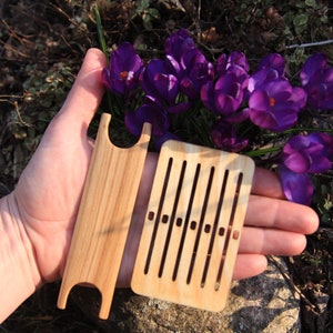 May include: Two wooden weaving tools, one rectangular and the other long and narrow, both with holes for threading yarn. The tools are held in a hand against a background of purple crocus flowers.