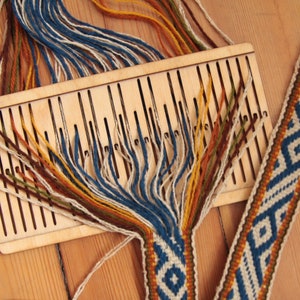 Op de afbeelding: Een houten weefgetouw met meerdere strengen garen in verschillende kleuren, waaronder blauw, geel, bruin en wit, die worden geweven tot een gepatroneerd weefsel. Het weefsel is een blauw, wit en bruin geometrisch ontwerp.