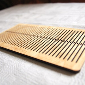 May include: A wooden loom with a dark brown border and the text "The Weaving Room" engraved on the bottom. The loom has a series of parallel slots for weaving yarn.
