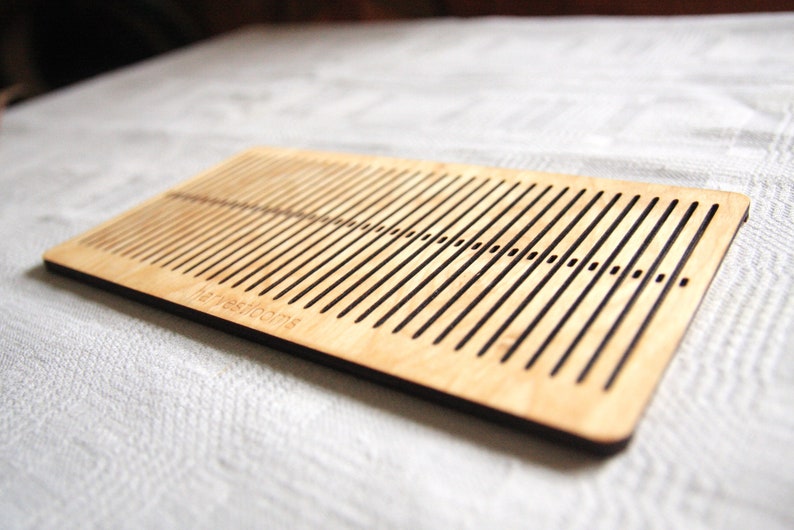 May include: A wooden loom with a dark brown border and the text "The Weaving Room" engraved on the bottom. The loom has a series of parallel slots for weaving yarn.