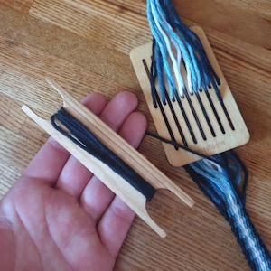 Peut inclure: Deux outils de tissage en bois avec du fil noir et bleu. L'un est une navette avec du fil noir, l'autre est un peigne à tisser avec du fil bleu, blanc et noir. Les outils sont sur une surface en bois.
