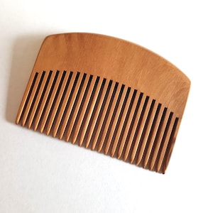May include: A wooden comb with a curved top and closely spaced teeth. The comb is made of light brown wood and is set against a white background. The comb's teeth are evenly spaced and run the length of the comb.