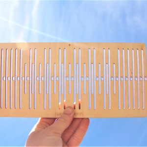 Op de afbeelding: Een houten weefgetouw met een lichtbruine afwerking en een patroon van verticale lijnen. Het weefgetouw wordt in een hand gehouden met de tekst "Yarn Rooms" zichtbaar aan de onderkant.