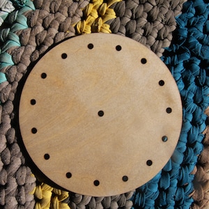 May include: A round, unfinished wooden craft piece with evenly spaced holes around the perimeter and one in the center. The wood has a light, natural color. The background is a colorful, textured rug with blue, green, brown, and yellow stripes.