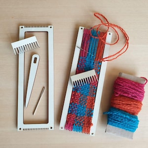 Op de afbeelding: Een ambachtelijke weefset met een rechthoekig frame, kam, naald en garen. Het geweven stuk heeft een kleurrijk patroon van blauwe, roze en oranje draden. Een spoel met roze, oranje en blauw garen is ook inbegrepen.