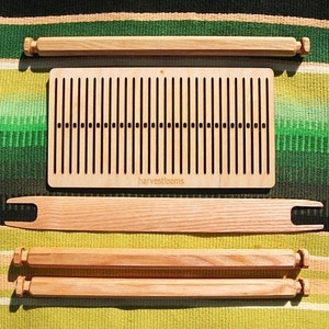 May include: A wooden loom with a natural finish. The loom is disassembled and laid out on a striped green and yellow blanket. The loom includes a weaving board with a label that reads "harvestlooms", two sets of wooden dowels, and a wooden shuttle.