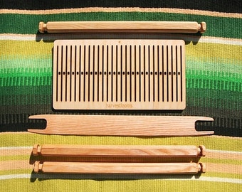 May include: A wooden loom with a natural finish. The loom is disassembled and laid out on a striped green and yellow blanket. The loom includes a weaving board with a label that reads "harvestlooms", two sets of wooden dowels, and a wooden shuttle.