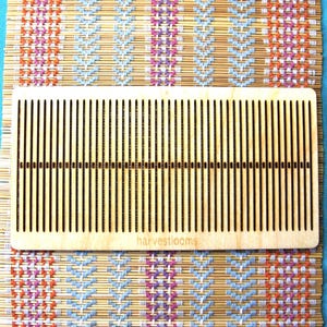 Peut inclure: Un peigne à tisser en bois avec des dents rapprochées, posé sur un tapis tissé avec un motif géométrique coloré. Le peigne porte l'inscription "harvestlooms". Le fond est un tissu turquoise uni.