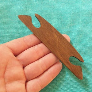 Op de afbeelding: Een bruine houten weefspoel, een hulpmiddel voor textielbewerking. De spoel heeft een langwerpige, slanke vorm met gebogen uiteinden en een gladde afwerking. Het wordt in een hand gehouden tegen een turquoise achtergrond.