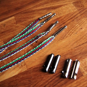 May include: A set of shoelaces featuring a woven design with red, green, and blue stripes, and silver aglets. The shoelaces are presented on a wooden surface, with several silver aglets. The shoelaces are new and unused.