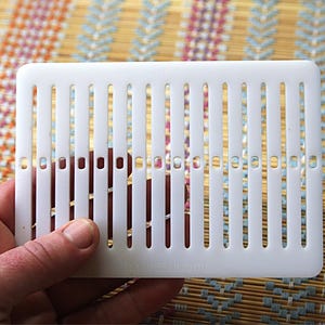 Op de afbeelding: Een witte, rechthoekige kunststof keukentool met verticale sleuven. De tool wordt in een hand gehouden en heeft de tekst "freshlooms" erop gedrukt. De achtergrond is een kleurrijke geweven mat.