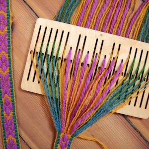 May include: A close-up of a wooden loom with colorful yarn threads woven through the slots. The loom is on a wooden surface. The yarn is green, yellow, and pink.