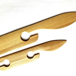 Puede incluir: Dos herramientas de tejido de madera con agujeros para enhebrar hilo. Las herramientas están hechas de madera marrón claro y tienen un acabado liso y pulido.