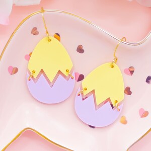 May include: A pair of yellow and purple acrylic earrings shaped like cracked Easter eggs. The earrings are hanging from gold hoops.