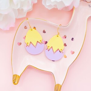 May include: A pair of yellow and purple egg-shaped earrings with gold hooks. The earrings are on a pink plate with gold trim.