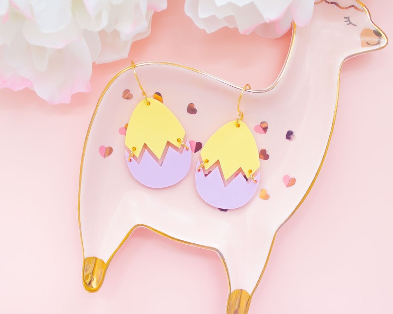 May include: A pair of yellow and purple egg-shaped earrings with gold hooks. The earrings are on a pink plate with gold trim.