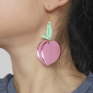 Pu&ograve; includere: Un paio di orecchini rosa a forma di pesca con un dettaglio a foglia verde. Gli orecchini sono in acrilico e hanno una finitura lucida.