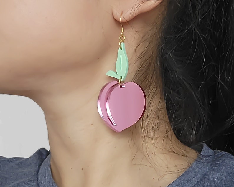Pu&ograve; includere: Un paio di orecchini rosa a forma di pesca con un dettaglio a foglia verde. Gli orecchini sono in acrilico e hanno una finitura lucida.