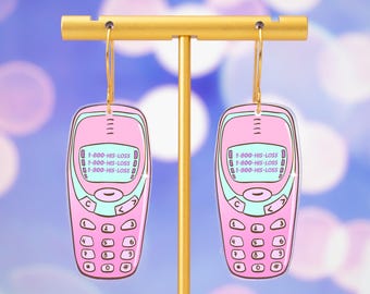 May include: Pink acrylic earrings shaped like retro cell phones, with a teal trim and gold hoops. The phone displays the text "1-800-HIS-LOSS". The earrings are displayed on a gold stand against a blurred background.