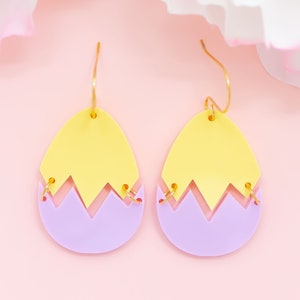 May include: A pair of yellow and purple acrylic earrings shaped like cracked eggs. The earrings have gold hooks.