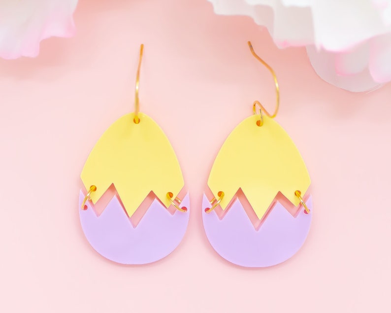 May include: A pair of yellow and purple acrylic earrings shaped like cracked eggs. The earrings have gold hooks.
