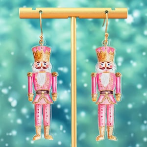 May include: Pink and gold nutcracker earrings. The nutcracker is wearing a pink and gold uniform with a gold crown. The earrings are hanging from a gold stand.
