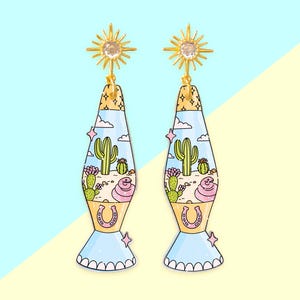 May include: A pair of gold-colored earrings with a sunburst design at the top and a lava lamp shape. The lava lamp design features a desert scene with cacti, clouds, and a horseshoe. The earrings are set against a light blue and yellow background.