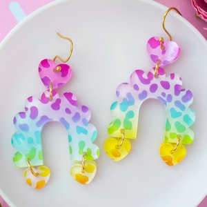 May include: Rainbow ombre acrylic earrings with a leopard print design. The earrings are shaped like an arch with a heart dangling from the bottom.