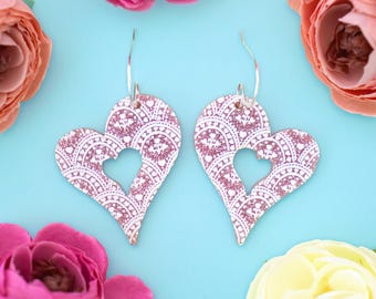 May include: A pair of pink heart-shaped earrings with a white filigree design and a silver hook. The earrings have a cutout heart shape in the center.