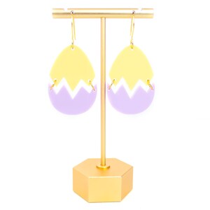 May include: A pair of yellow and purple egg-shaped earrings with a gold hook. The earrings are on a gold stand.