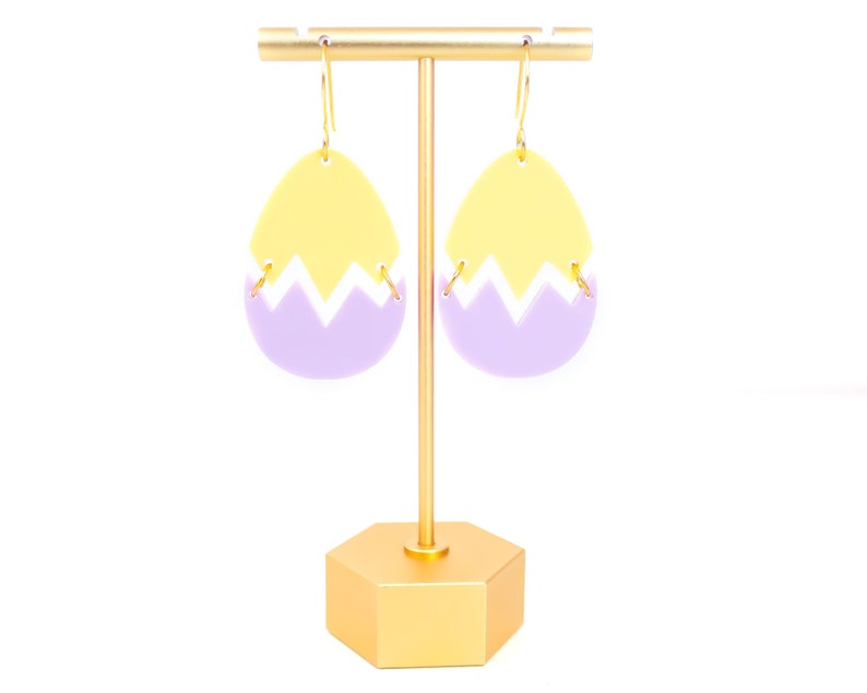 May include: A pair of yellow and purple egg-shaped earrings with a gold hook. The earrings are on a gold stand.