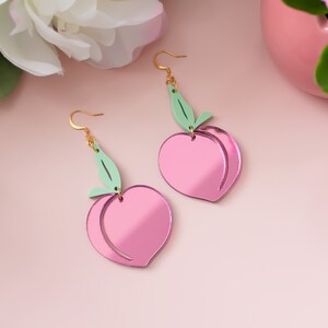 Pu&ograve; includere: Un paio di orecchini rosa a forma di pesca con accenti di foglie verdi. Gli orecchini sono in acrilico e hanno una finitura a specchio.