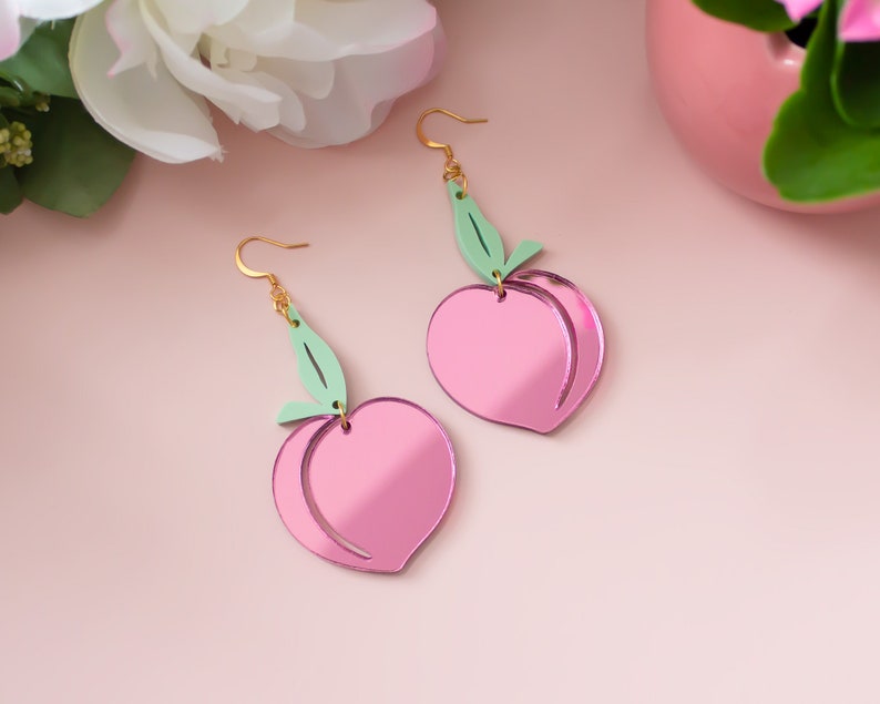 Pu&ograve; includere: Un paio di orecchini rosa a forma di pesca con accenti di foglie verdi. Gli orecchini sono in acrilico e hanno una finitura a specchio.
