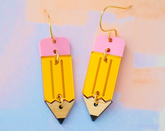 May include: A pair of yellow and pink pencil earrings with gold accents. The earrings are made of acrylic and have a wood-like finish at the bottom.