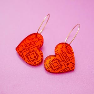 May include: A pair of heart-shaped earrings with a circuit board design. The earrings are a transparent orange color with a dark brown pattern. They hang from thin, gold-colored hoops. The background is a solid pink.