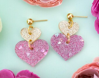 May include: A pair of gold and pink heart-shaped earrings. The earrings are made of glitter resin and have a layered design. The top heart is gold and the bottom heart is pink.
