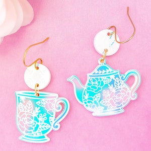 May include: A pair of iridescent blue and white teapot and teacup earrings. The teapot and teacup are decorated with a white floral design. The earrings are on a gold hook.
