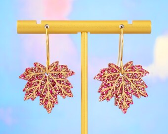 Op de afbeelding: Een paar gouden oorbellen in de vorm van esdoornbladeren met roze glitter. De oorbellen hangen aan een gouden standaard.