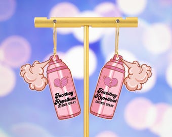May include: A pair of pink spray bottle earrings with the text "Fuckboy Repellent DEFENSE SPRAY" and a broken heart graphic. The earrings are hanging from a gold-colored display stand against a blurred blue background.