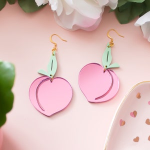 Pu&ograve; includere: Un paio di orecchini rosa a forma di pesca con foglie verdi. Gli orecchini sono in acrilico e hanno una finitura lucida.