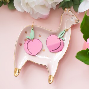 Pu&ograve; includere: Un paio di orecchini a forma di pesca con foglie verdi, esposti su un piatto in ceramica bianca a forma di lama con dettagli dorati. Gli orecchini sono rosa e hanno una finitura lucida.