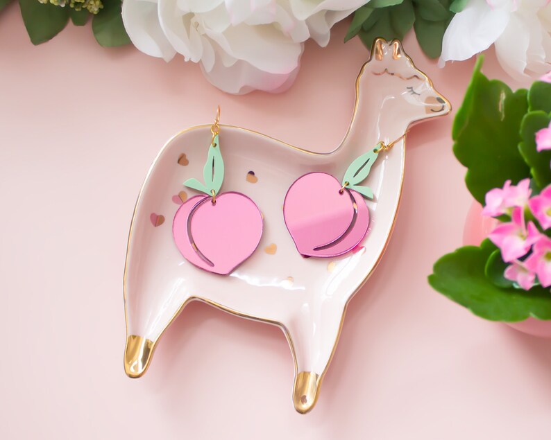 Pu&ograve; includere: Un paio di orecchini a forma di pesca con foglie verdi, esposti su un piatto in ceramica bianca a forma di lama con dettagli dorati. Gli orecchini sono rosa e hanno una finitura lucida.