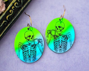 May include: A pair of iridescent green and blue earrings featuring a skeleton holding a coffee cup. The earrings are made of acrylic and have a gold-tone hook closure.