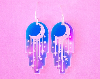 May include: A pair of celestial-themed earrings. Each earring features a blue and purple gradient design with a crescent moon and stars. The earrings have a layered, geometric shape and dangle from silver-colored hooks.