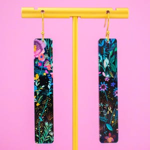 May include: A pair of long, rectangular earrings with a floral pattern. The earrings are black with colourful flowers and leaves. The earrings are hanging from a gold stand.