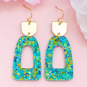 Peut inclure: Une paire de boucles d'oreilles imposantes bleu turquoise avec des accents dorés. Les boucles d'oreilles sont en résine et ont une finition scintillante. Les boucles d'oreilles sont en forme d'arche avec une petite arche dorée en haut.